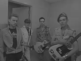 Four men standing in a room, one of the far left holding a bass guitar, and to his right holding an electric guitar
