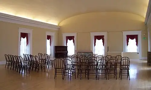 large, open meeting room with a curved ceiling, curtained windows, several rows of wooden chairs, and a piano in the corner between two of the windows