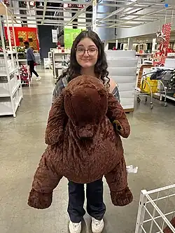 A young woman is holding the plush bear inside an IKEA store. The bear is about a metre tall
