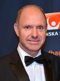 A man wearing a black suit and bow-tie is shown from the shoulders up