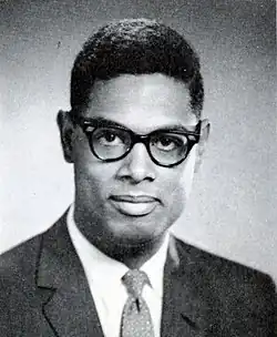 A dark-haired man, wearing glasses and a suit and tie, looks into the camera