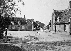 Large thatched cottage on right, man standing in front of pond on left, large thatched cottage in background with a small group of people and two horses standing nearby