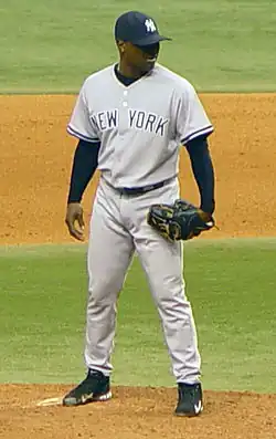 A dark-skinned man in a gray baseball uniform with "New York" across the chest in navy-blue letters and a navy-blue baseball cap standing on a dirt mound on a grass field and wearing a black baseball glove on his left hand