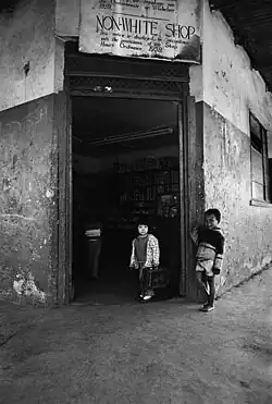 A black and white photo of two children in front of a store with a sign that says: "NON-WHITE SHOP This notice is displayed in accordance with the provisions of the Shop Hours Ordinance. 1959."