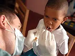 white man holding plastic tray with brown goop in it and sticking a small stick into a black boy's open mouth