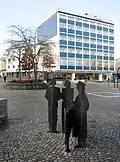 Six-story white and blue hotel building in a city centre with a red sign reading "Scandic". In the middle of the picture a white square, "Rutan" ("The square"), is painted on the cobblestone. In the foreground of the picture there are statues of three persons and a cow.
