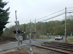 Train tracks going from lower right to mid-left cross, at grade, with metal plating around the tracks, a paved road going lower left to an intersection at a divided highway with a traffic light just above the tracks. Two railroad crossing signs with red-and-white-striped barriers in the up position are on either side of the tracks. A pine tree's branches protrude into the image from the left; across the divided highway are more woods, with trees beginning to show leaves, slightly muted under an overcast sky