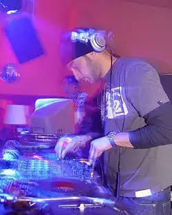 A DJ wearing headphones is performing using his turntables and other electronic equipment. A computer monitor is visible in the background.