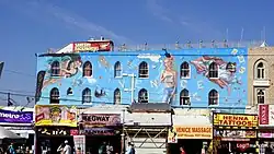 View of mural seen over small store front businesses. Mural has a woman whose thoughts appear as text in a cloud saying, "History is myth."