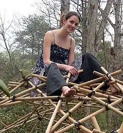 Portrait of Vi Hart, a young white person with long hair in a bun, sitting on top of a finished mathematical project, a polyhedron made of bamboo sticks and rope.