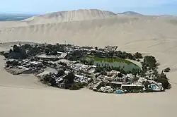 Small lagoon surrounded by palm trees and village buildings, nestled among tall sand dunes