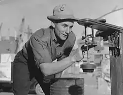 A smiling man in a short-sleeved shirt, dark pants, and a light-colored bucket hat examining a piece of mechanical equipment