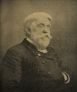 A white-haired man with a beard and mustache, facing left. He is wearing a white shirt, black vest, and black jacket