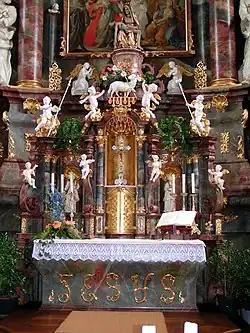 The altar and the reredos that rises behind it together are an example of German Baroque church fitting. They have polychrome marbled surfaces of pink and grey which match the columns of the church. The reredos is heavily architectonic with columns, niches, scrolls and pediments of different shapes which rise in stages to a plinth on which rests a Medieval statue of the Virgin Mary grieving over the body of Christ. On the level below this is a statue of the Lamb of God. In the niche beneath is a silver cross of Medieval design. Beneath this is a gilt metal sacrament cupboard. The central objects are surrounded by a profusion of shiny white stucco cherubs, angels and saints. Across the front of the marble altar is the name "Jesus" is elaborate capital letters. The altar is decked with a lace-edged cloth, flowers and a Bible.