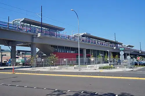 The elevated Wilburton station under construction in August 2022. The station has a concrete platform and a glass-enclosed waiting area. The station is surrounded by a chain-link fence and there's a parking lot nearby. The sky is clear and blue.