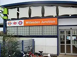 A white-panelled building with a rectangular, orange sign reading "Willesdon Junction" in white letters all under a light blue sky