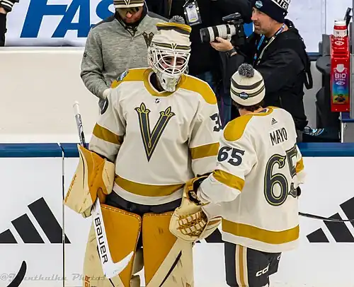 Two ice hockey players, a skater and a goaltender, in off-white jerseys with a grey "V" crest, gold shoulders/stripes, and grey equipment. Both are wearing off-white, grey, and gold toques. The skaters has his back to the camera, while the goaltender faces it.