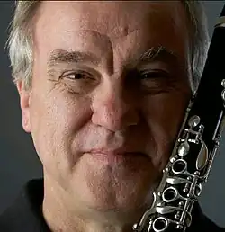 Colour publicity shot of a clean-shaven, smiling man with short, receding grey hair. He his holding a clarinet close to his face.