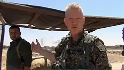 Michael Enright wearing army uniform, squinting and appearing to speak directly to camera, standing under makeshift shelter with smoke in the background and other armed men next to him