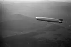 A black-and-white photograph from slightly above of the Graf Zeppelin, a large slim airship, flying from right to left against a low sun. The ground below is misty and consists of rolling hills, woods, and a meandering road which is catching the sun. The silvery airship is also reflecting the sun. It bears black swastikas on white circles on its vertical tail surfaces.